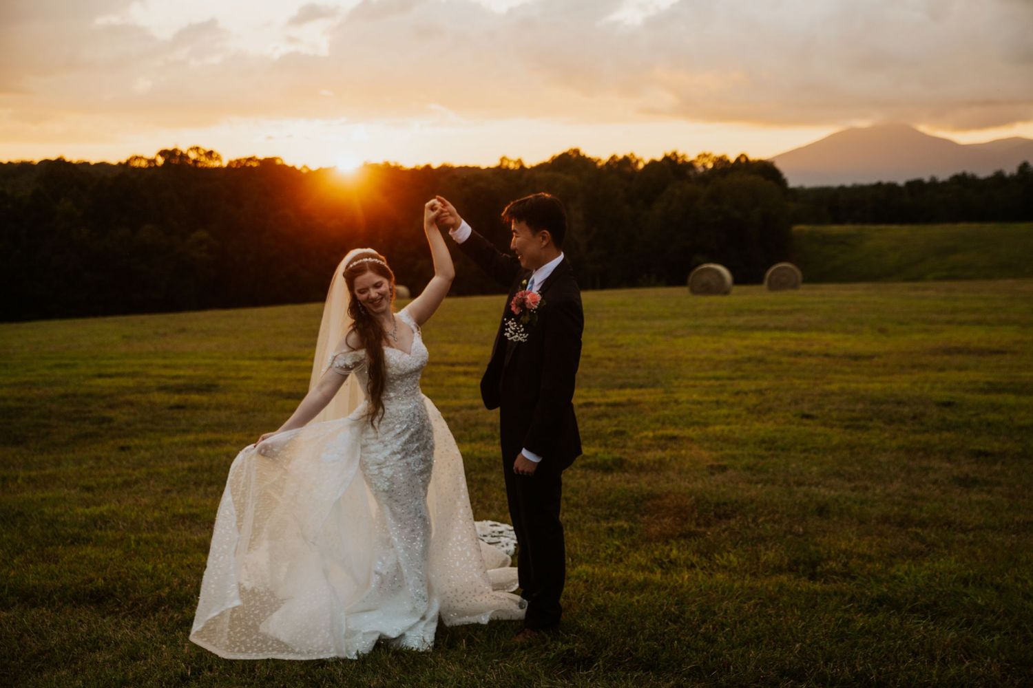 Bride and groom dance at golden sunset on lush green farm field with hay bales and mountains in background.
