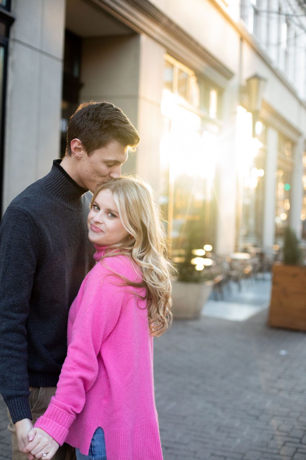 Couple in pink and black sweaters embracing on an urban sidewalk during sunset.