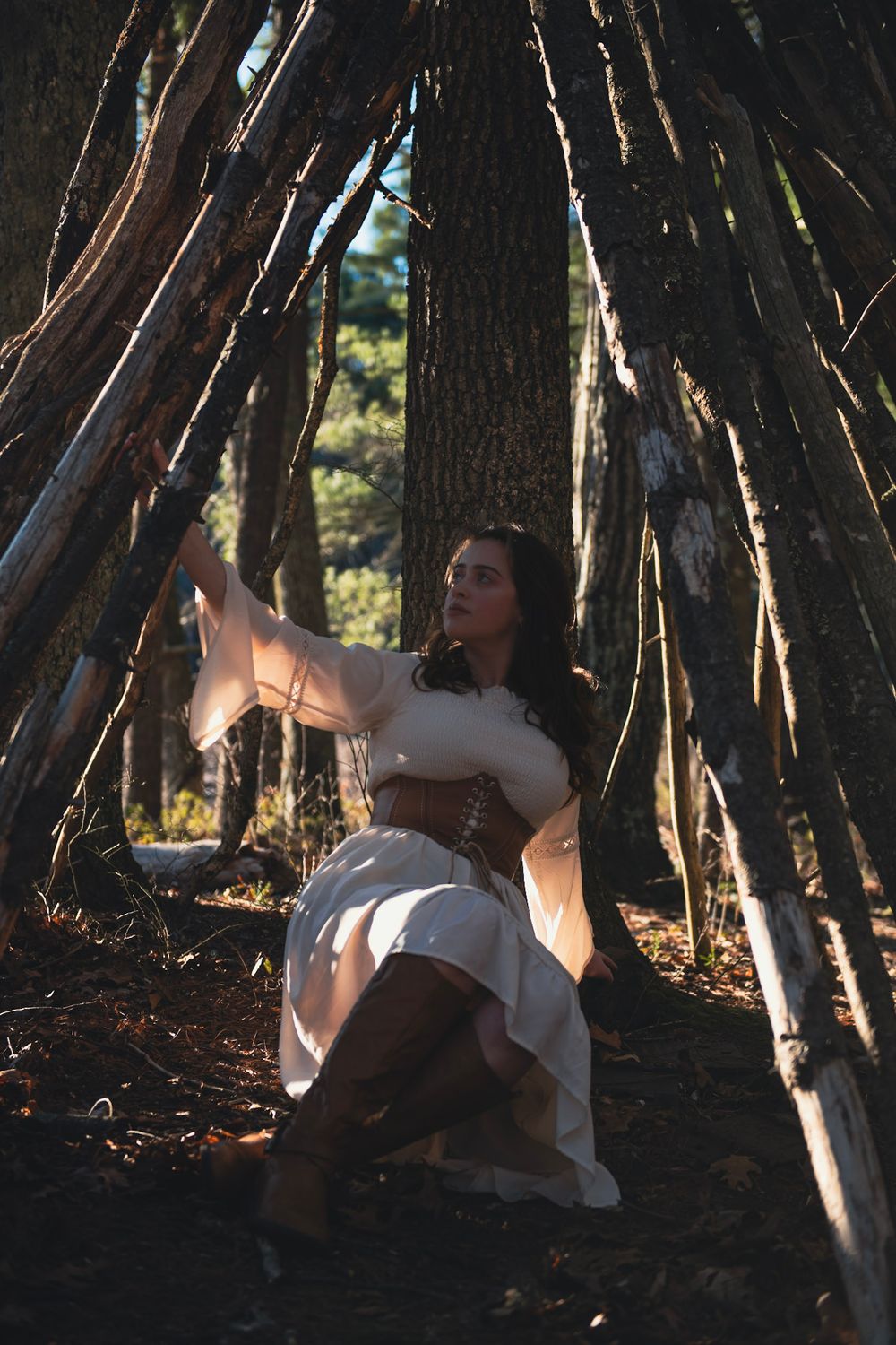 An ethereal figure in a white dress poses inside a teepee-like structure made of wooden sticks in a forest.