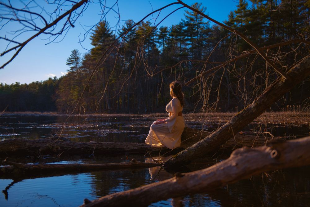 A figure in a white dress sits contemplatively on a fallen log by a serene lake at sunset.