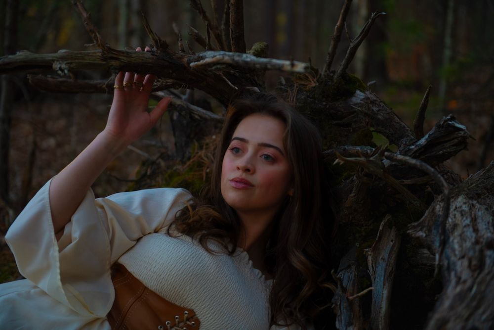 An artistic forest portrait shows someone in a white dress and brown corset among tree branches.