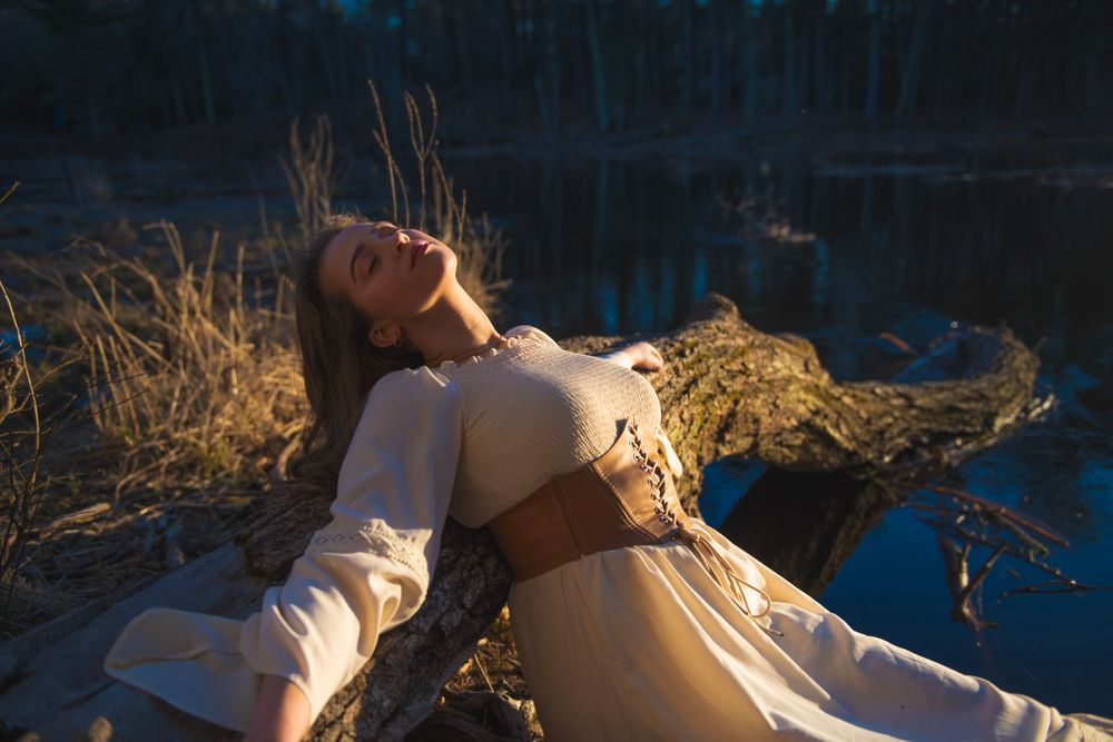 A sun-drenched portrait captures a reclining figure in white dress and brown corset by a lake.