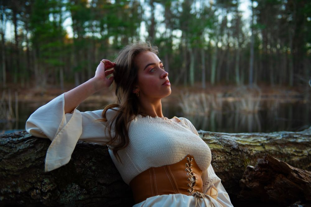 A dramatic outdoor portrait shows a figure in white dress and brown corset against a forested lake backdrop.