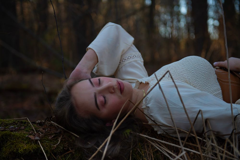 A dreamy portrait shows someone lying on mossy ground in a white dress during golden hour.
