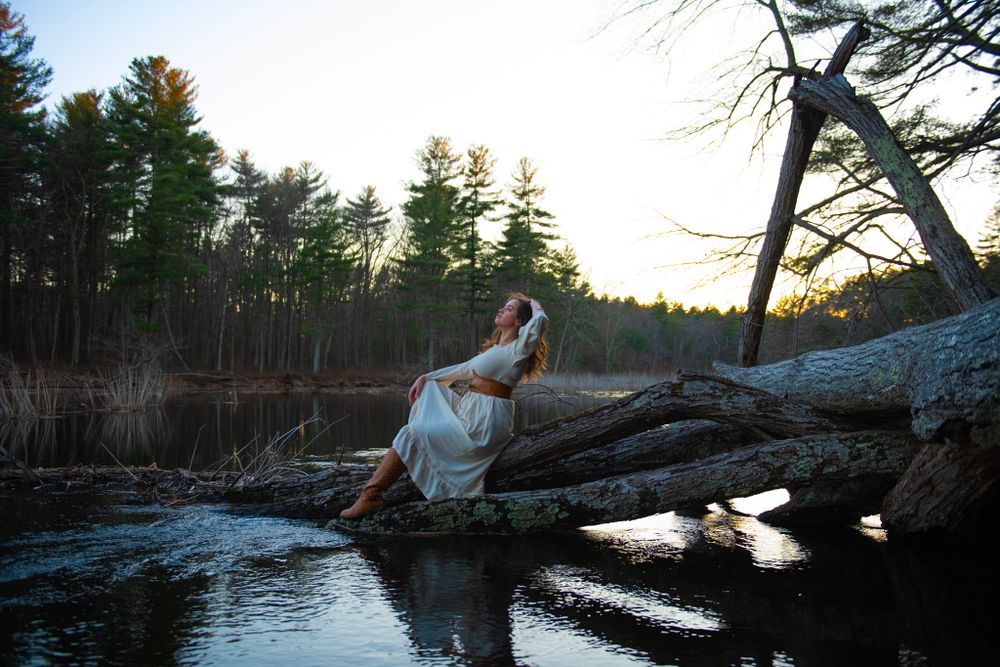 White-clad figure perches gracefully on fallen log over still water at sunset.