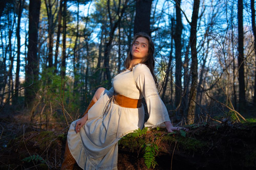 A figure in a flowing white dress with brown belt poses gracefully against a forested backdrop.