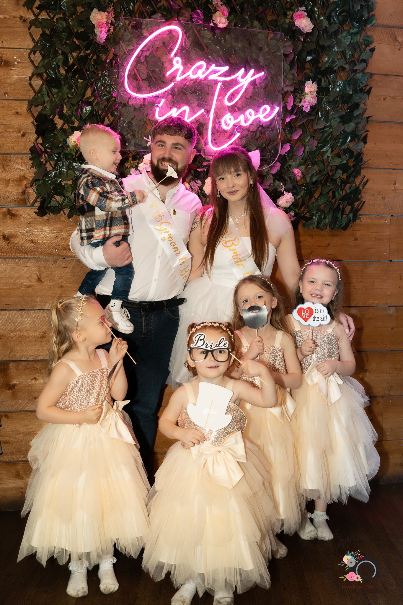 Group photo with pink neon 'Crazy in Love' sign and children in tulle dresses against wooden wall.