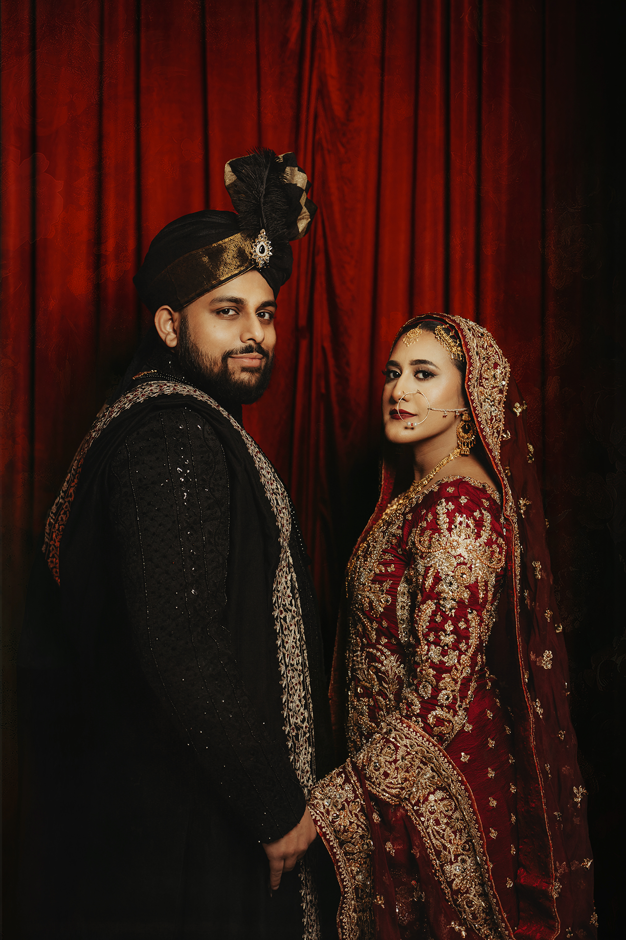 Muslim couple in elegant and royal wedding attire pose against a red curtain backdrop in dramatic lighting.