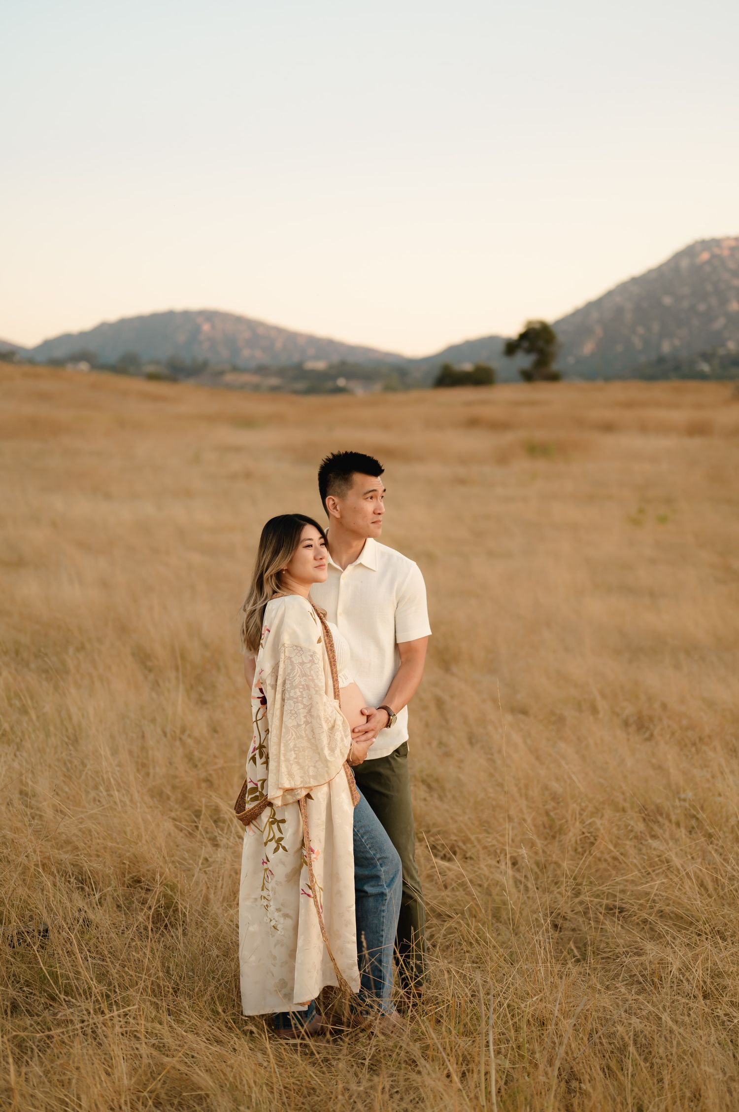 husband and wife hold pregnant belly during their Ojai maternity photo shoot in golden grassy field by Santa Barbara photographer