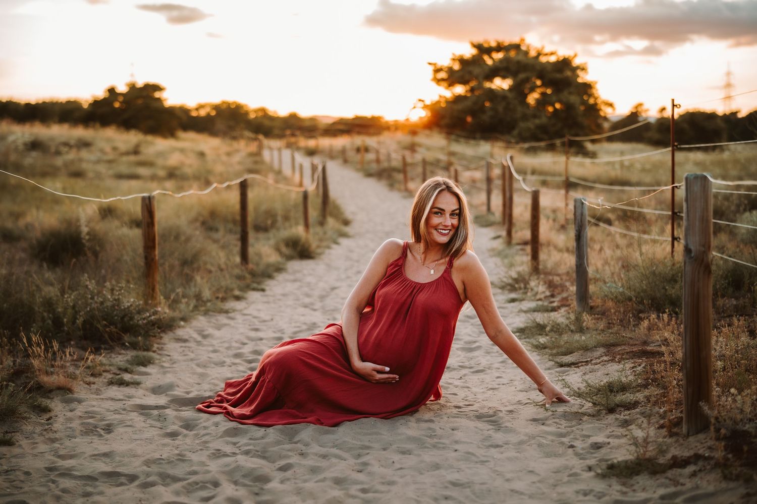 Woman in flowing red dress posing on sandy path during golden hour sunset.