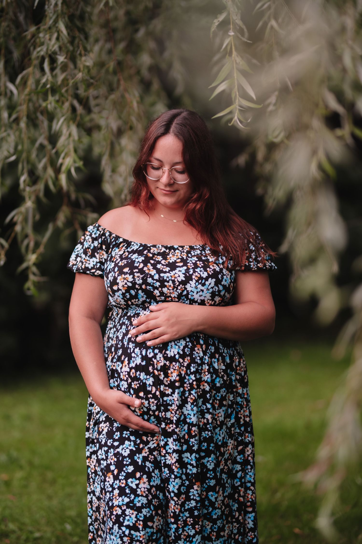 Expectant mother in floral dress smiling while standing under willow tree branches.