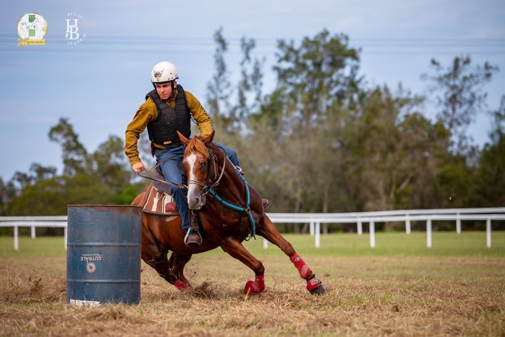 A horse and rider compete in barrel racing on a dirt track, making a tight turn around a blue barrel in sunlight.