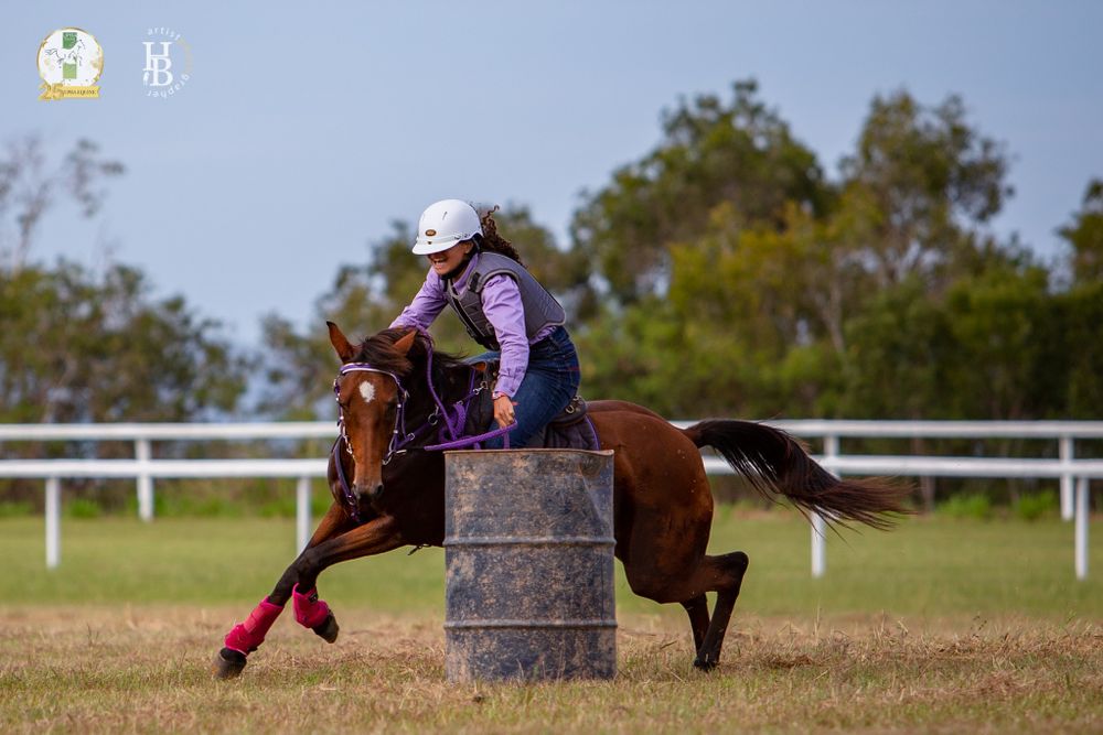 A rider on a chestnut horse performs barrel racing on a dirt arena with green trees in the background.