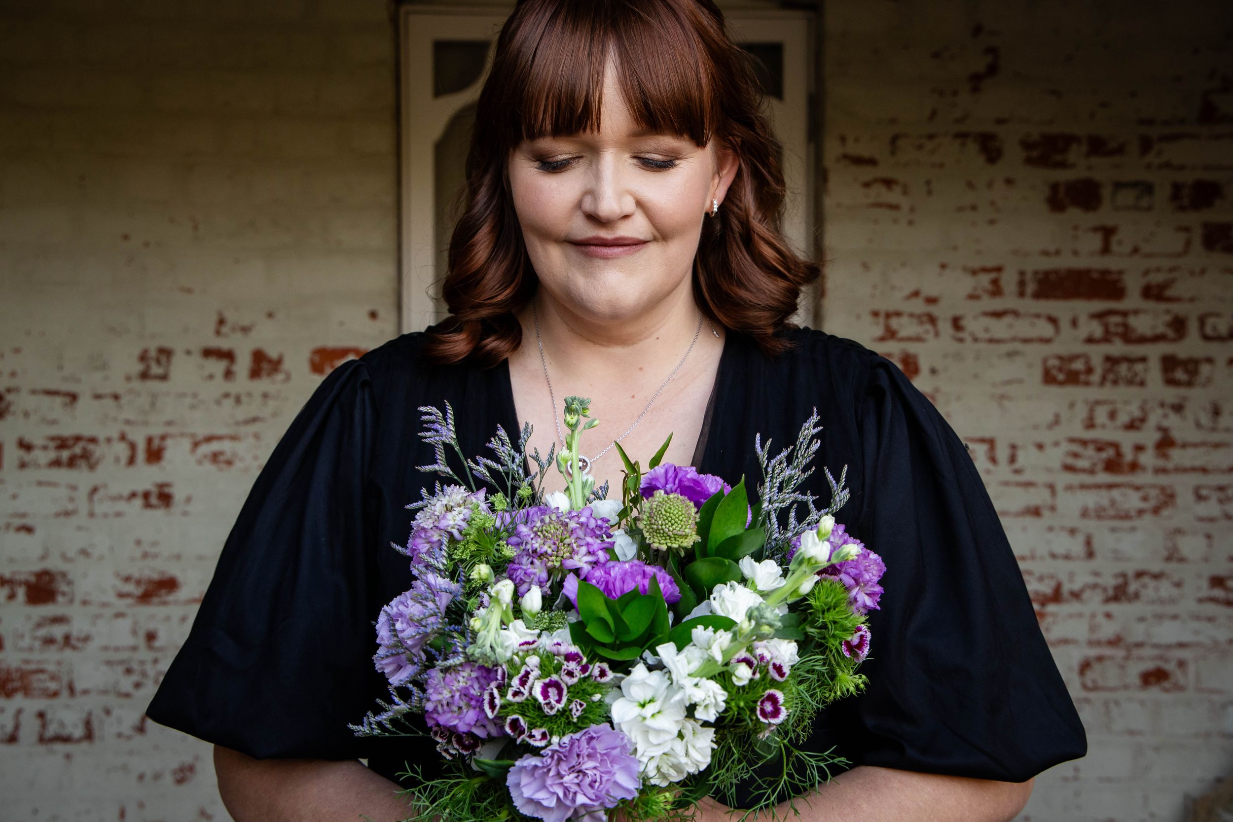 Person holding beautiful purple and white floral bouquet against brick wall.
