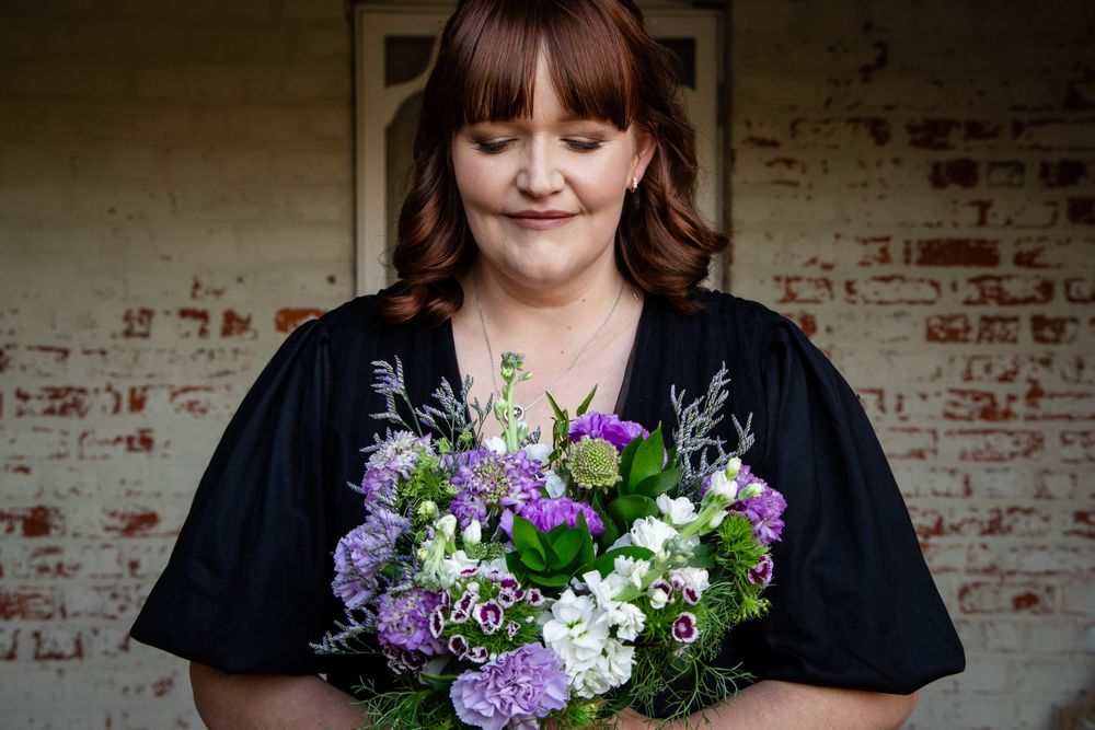 Person holding beautiful purple and white floral bouquet against brick wall.