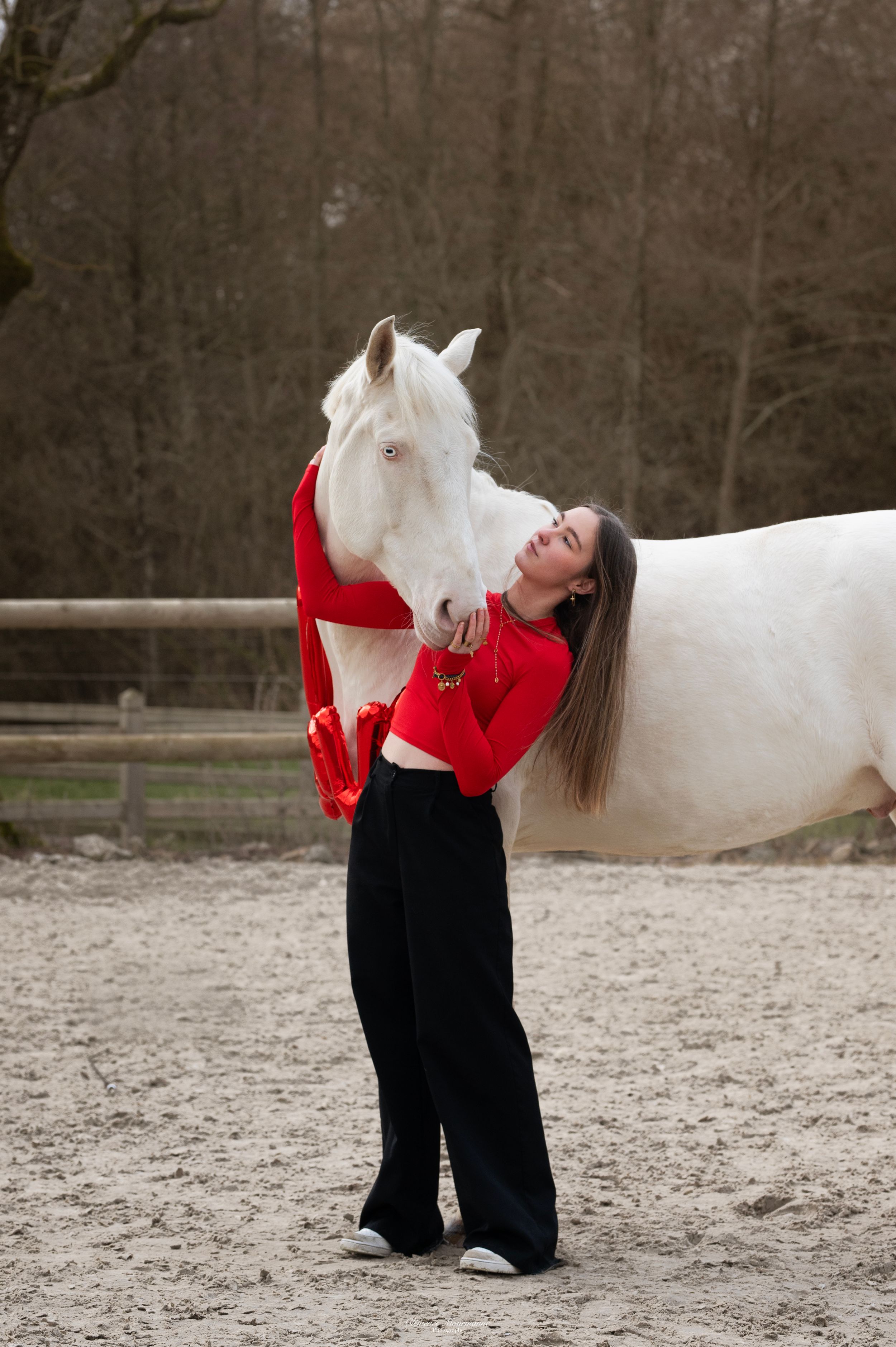 A white horse and person in red and black clothing share a tender moment in an outdoor arena.