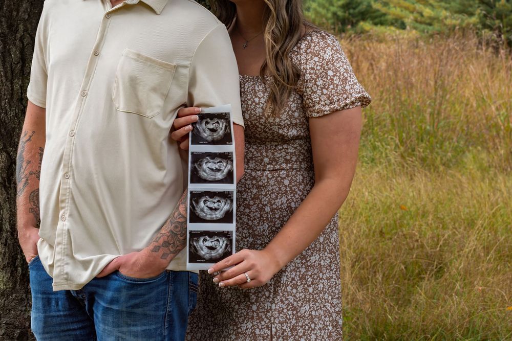 A couple in a grassy field holds up ultrasound photos while sharing their pregnancy announcement.