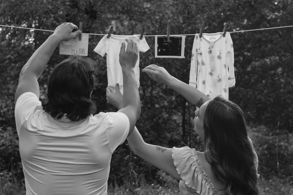 Black and white photo of two people hanging baby clothes on a clothesline outdoors.