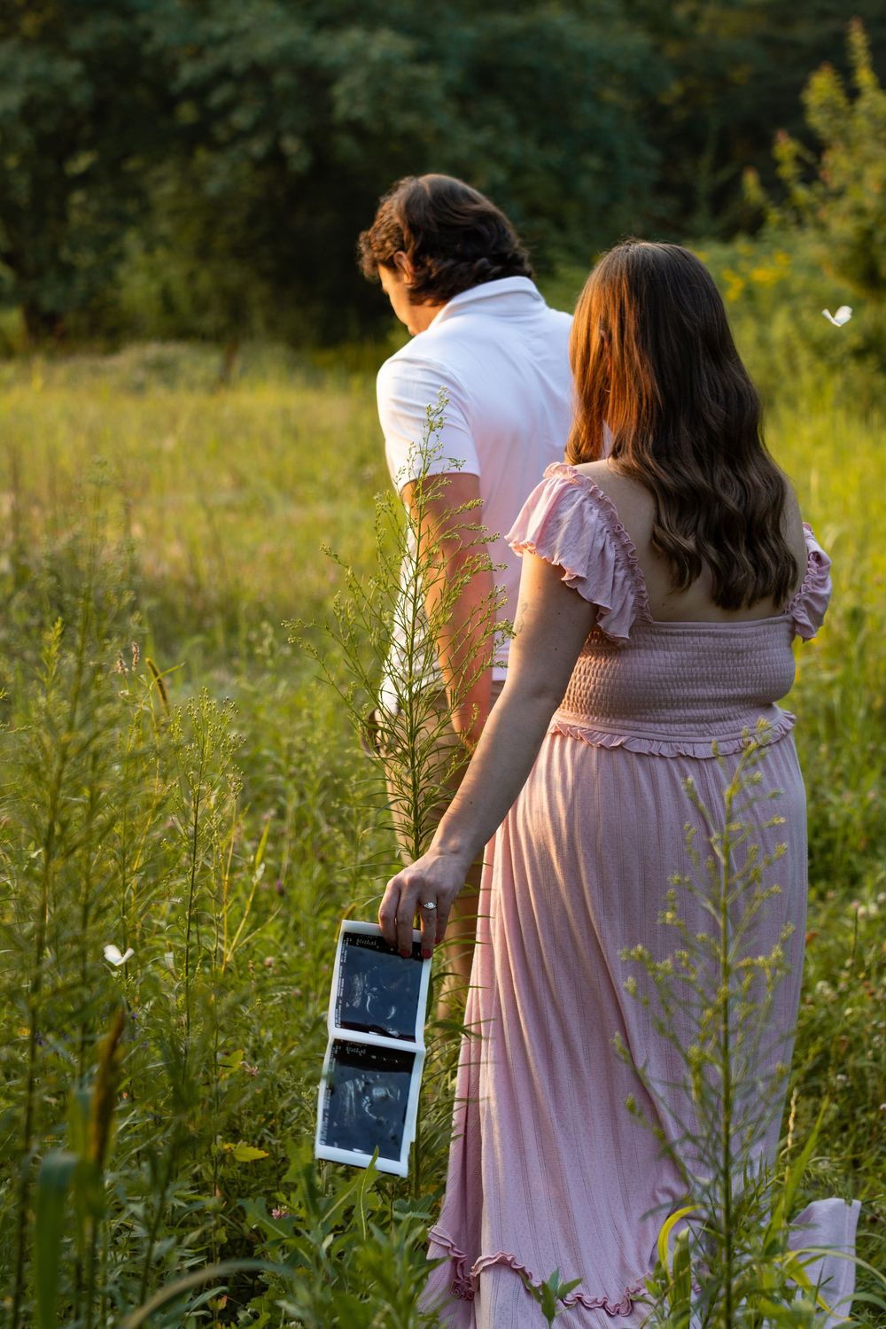 A couple standing in a wildflower meadow at sunset holding an ultrasound photo in a romantic maternity moment.