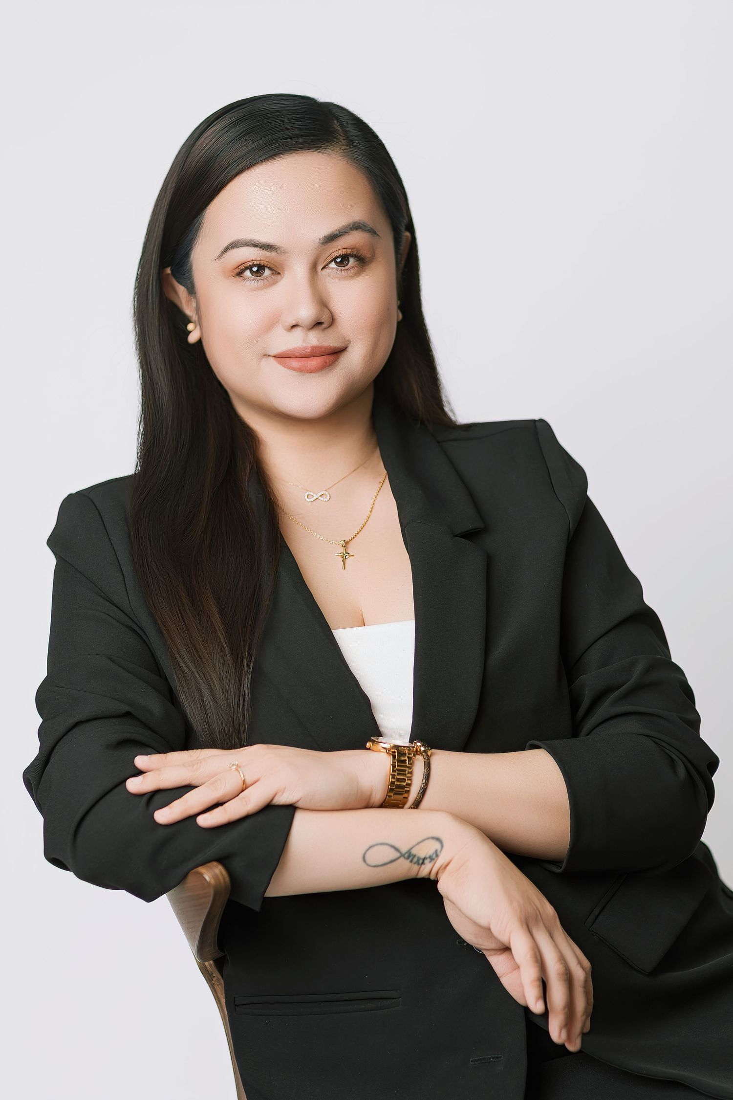 Professional headshot of a person in a black blazer and white top against a light background, with long dark hair and jewelry.