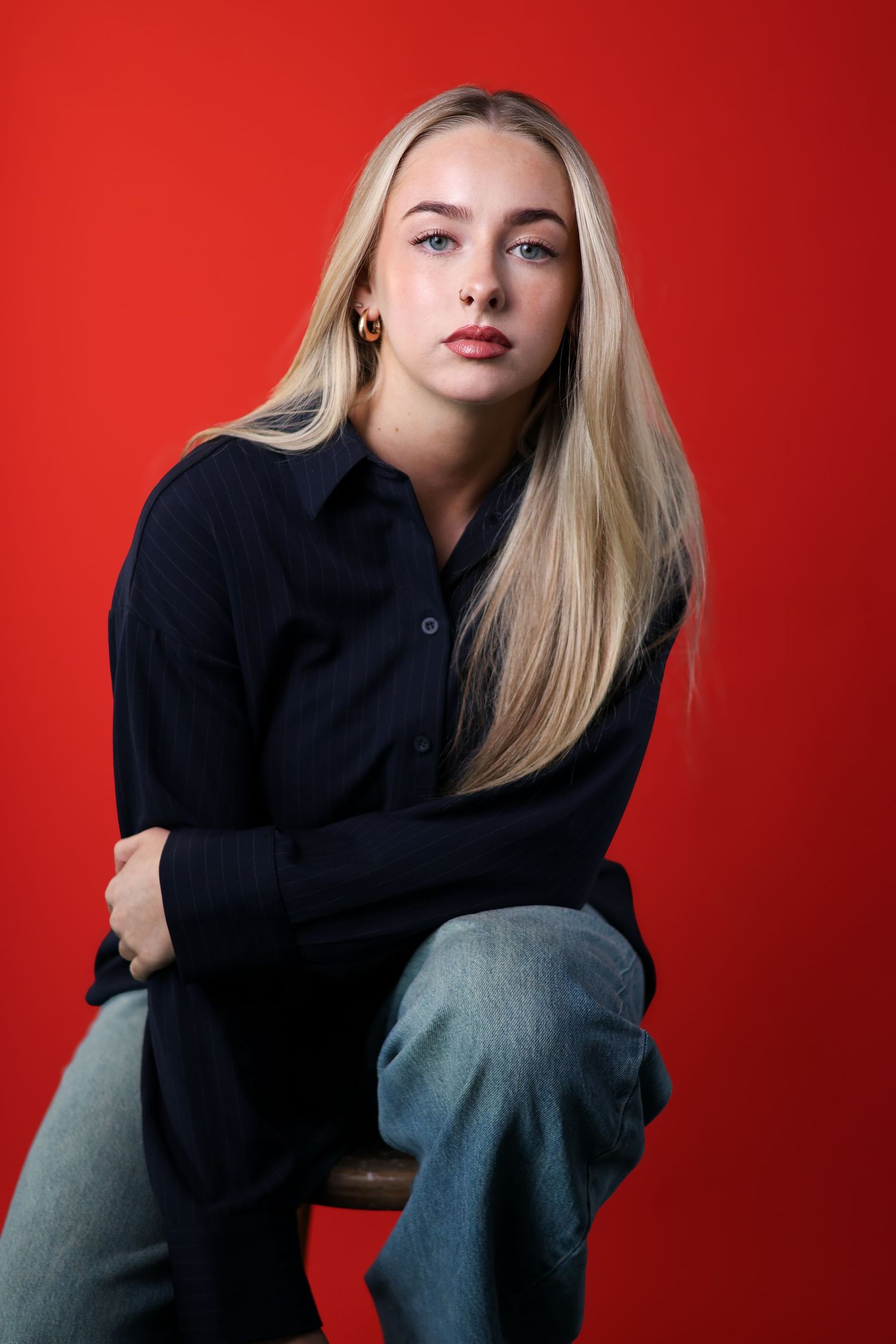 Young woman in pink jacket leans thoughtfully on wooden chair in studio setting.
