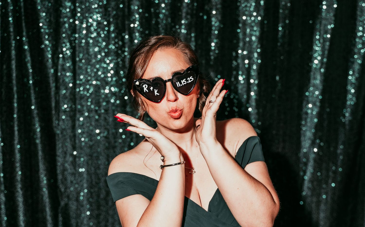 A person wearing sunglasses poses playfully against a sparkly green sequin backdrop.