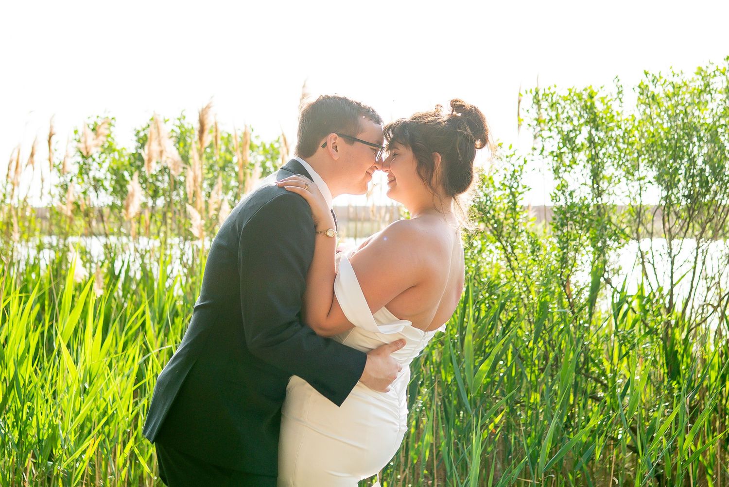 A romantic wedding couple embraces among tall green reeds at sunset beside a lake.