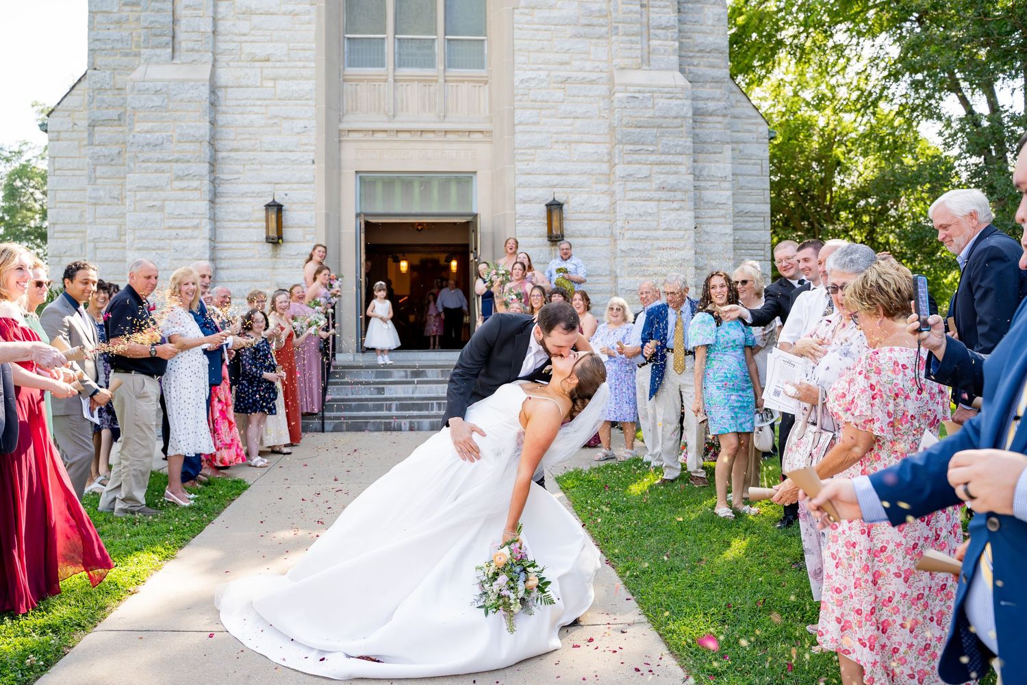 Celebratory moment with wedding party outside a stone church as crowd throws petals during summer wedding ceremony.