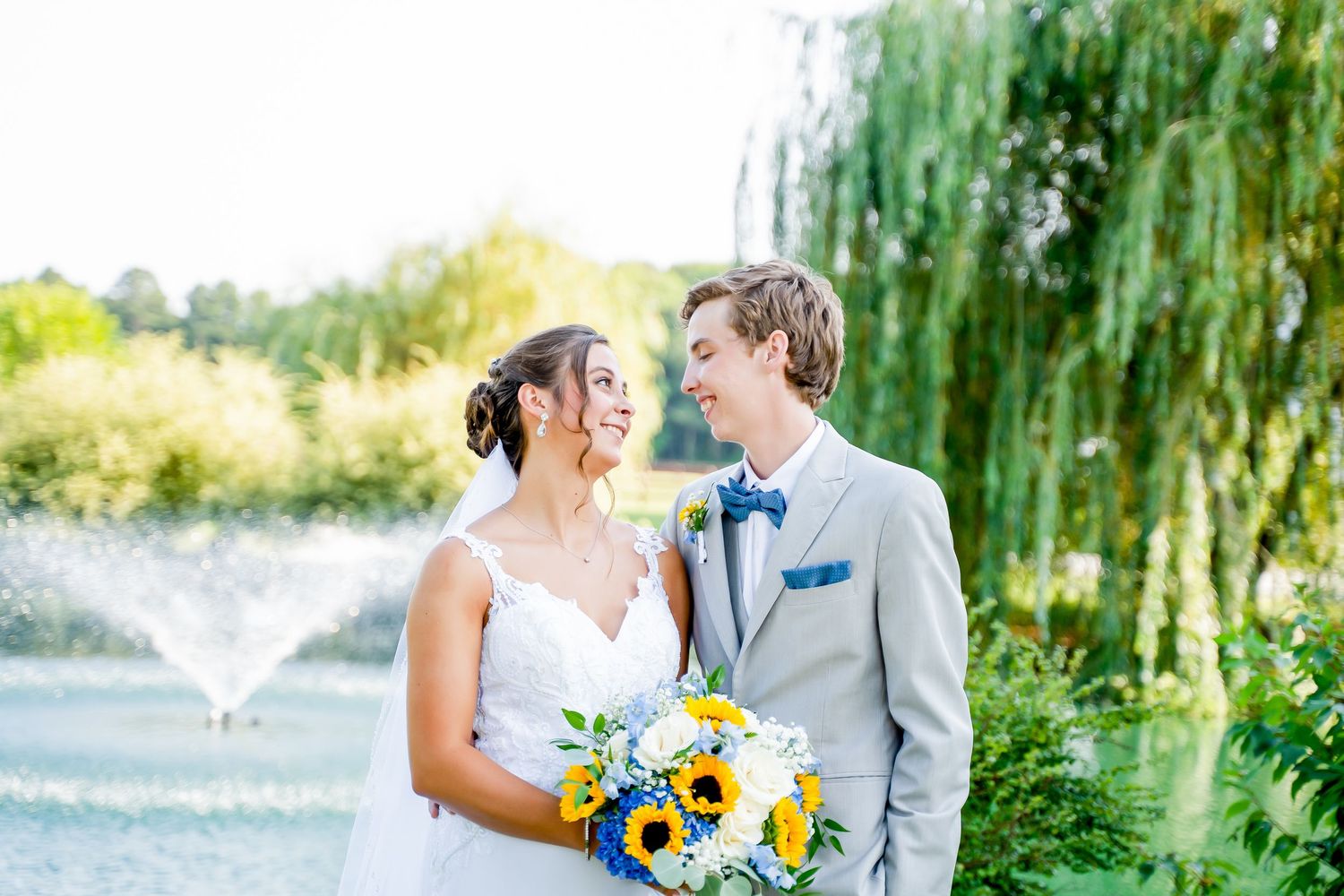 Two people in wedding attire share a loving moment under a weeping willow tree with sunflowers in the bouquet.