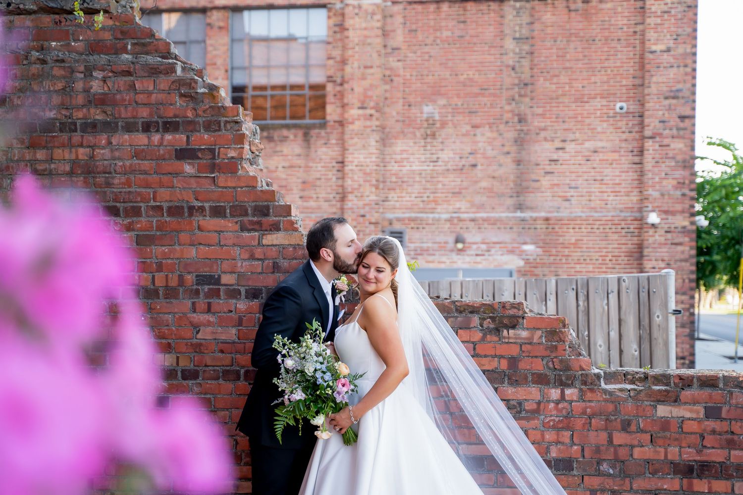 A romantic wedding moment captured against a rustic brick wall with pink flowers in the foreground.