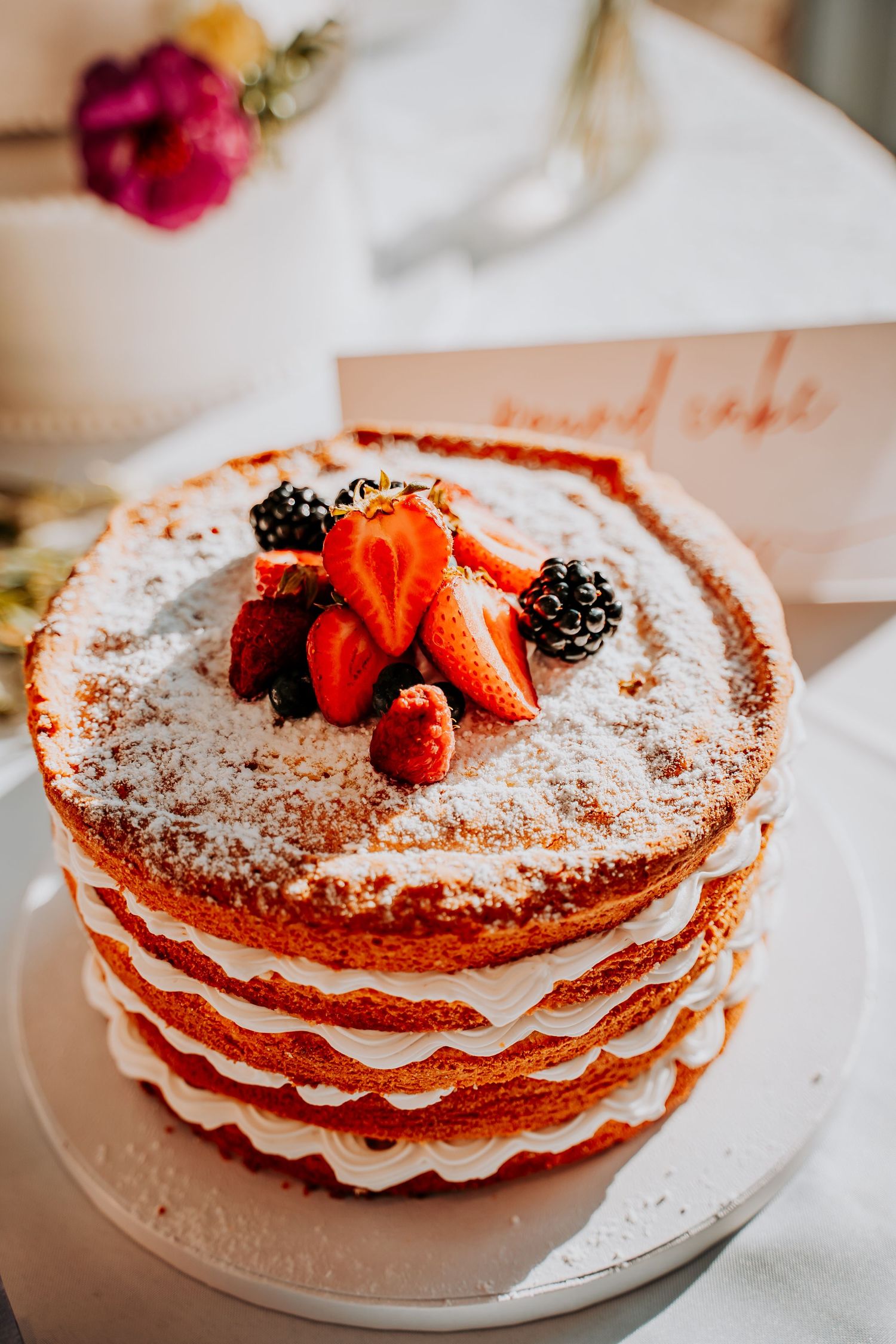 Multi-layered vanilla cake topped with fresh strawberries, blackberries and powdered sugar on a white plate.