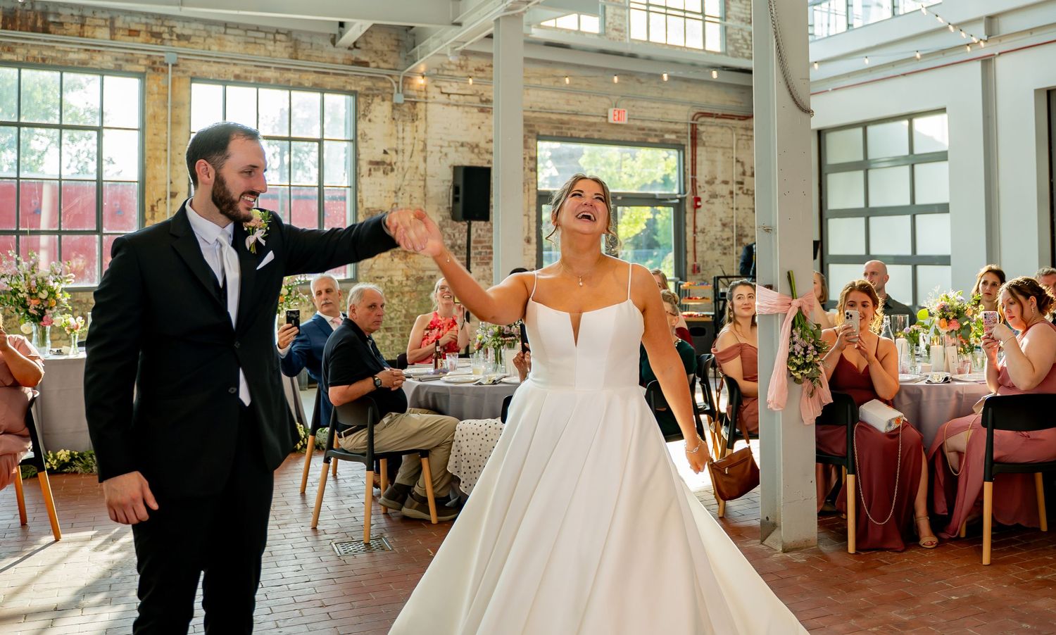 A couple in elegant wedding attire dances together during their first dance in a bright sunlit venue.
