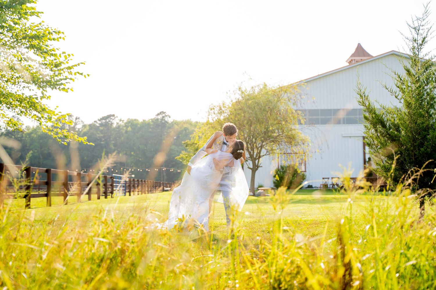 A romantic moment captured in a sunlit meadow outside a white barn as sunbeams filter through the golden grass.