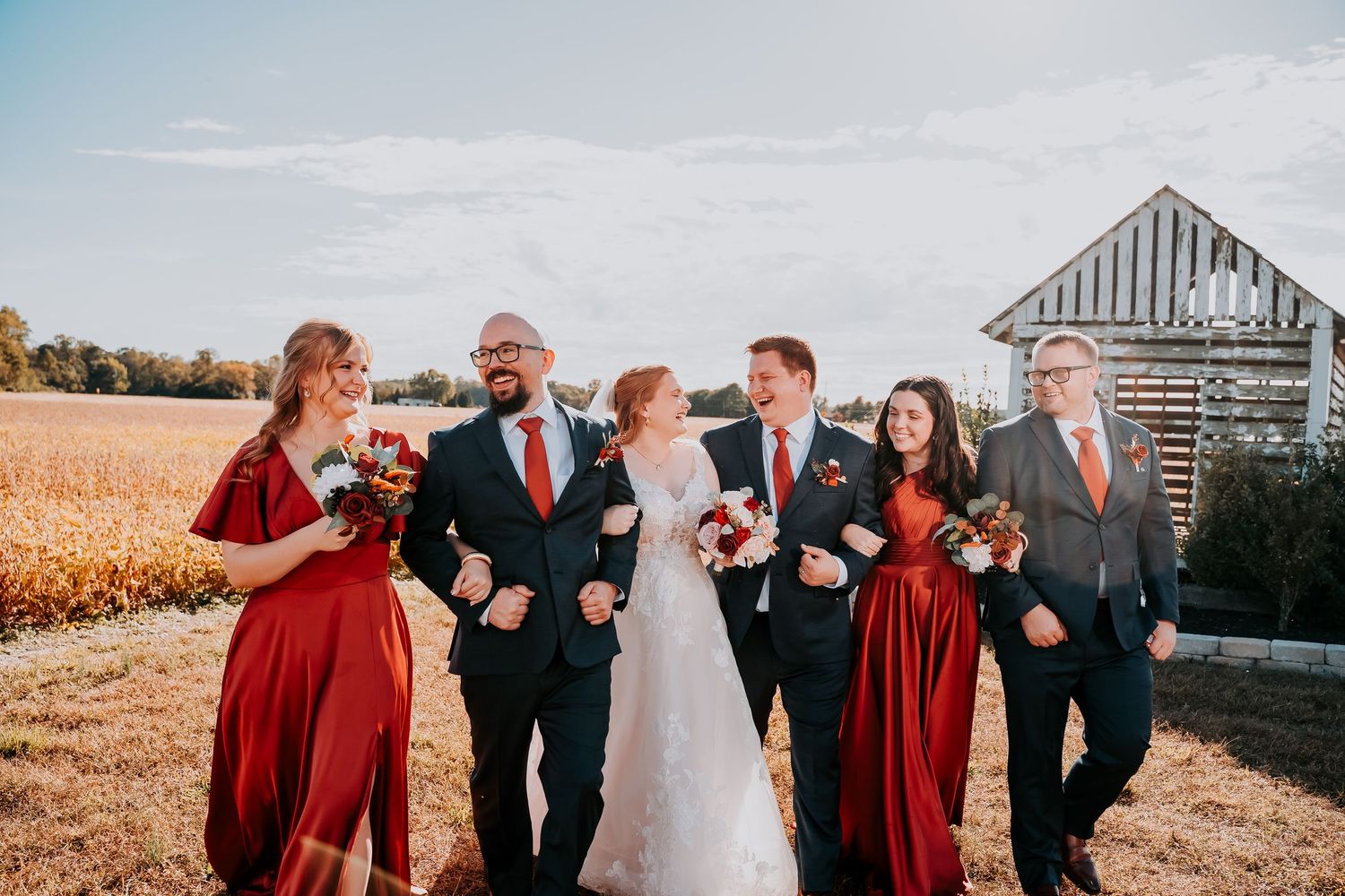Wedding party members laugh and pose together in front of rustic barn and cornfield at sunset.