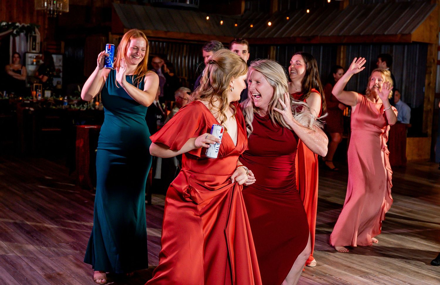 Wedding guests in red and pink dresses dance together on a dark wooden dance floor at an evening reception.