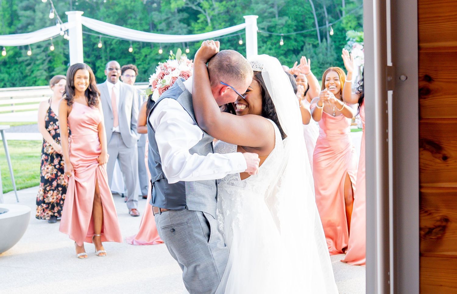 Wedding party in pink and gray attire poses together in front of wooden cabin during outdoor celebration.