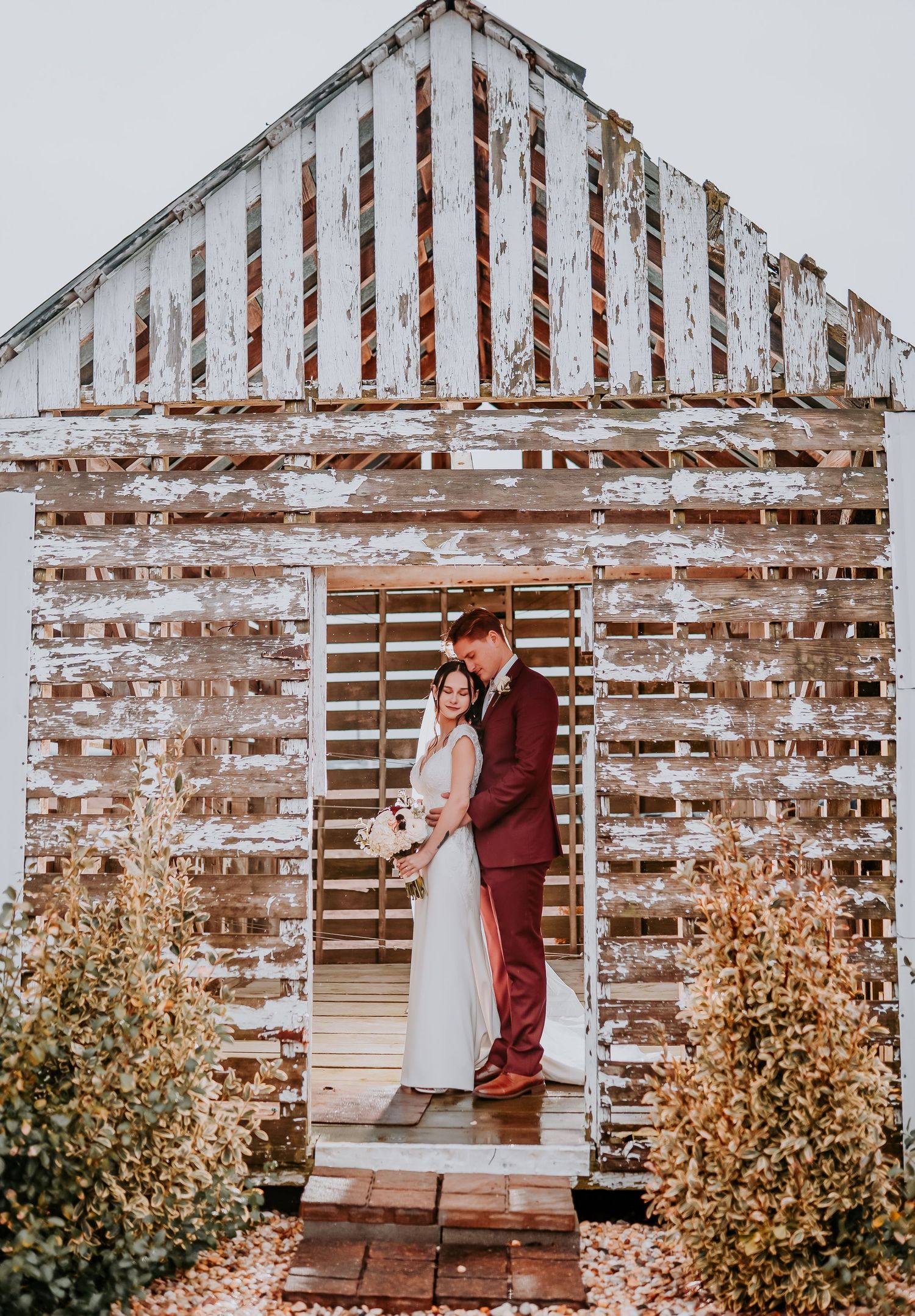 A rustic white wooden barn with peeling paint serves as a backdrop for a couple in burgundy and white attire.