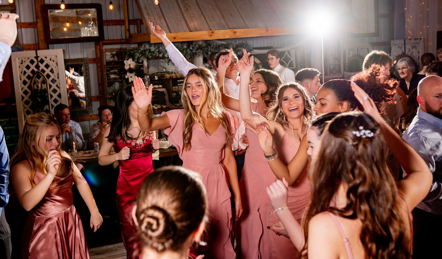 A crowd of wedding guests dance energetically on a dark dance floor illuminated by bright reception lighting.