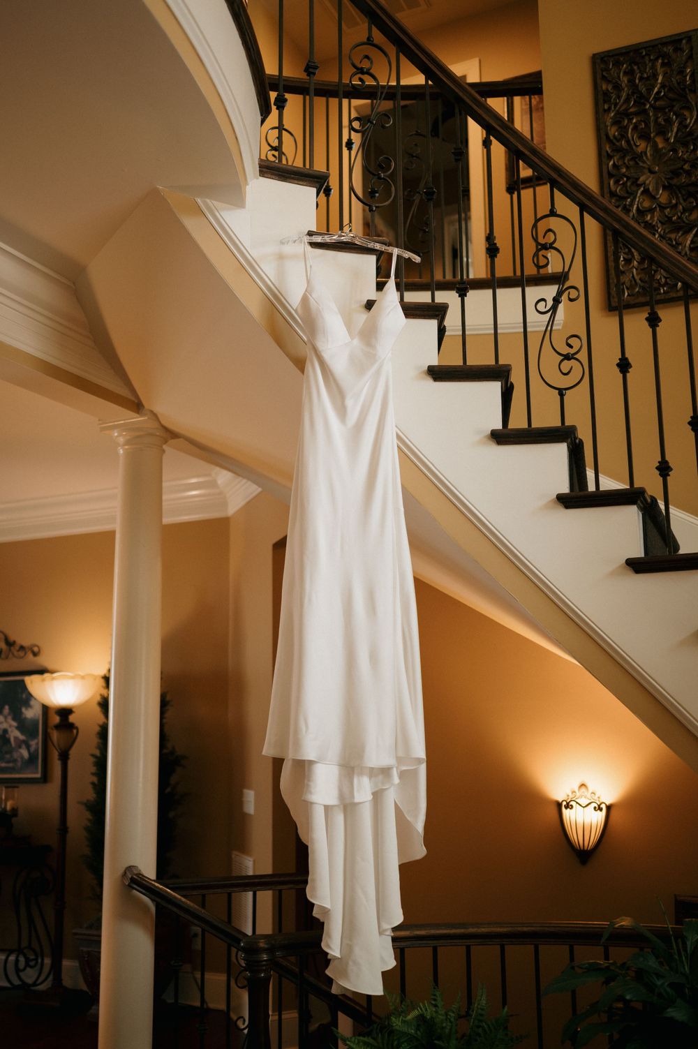 An elegant white wedding dress hanging on a curved staircase with wrought iron railings.