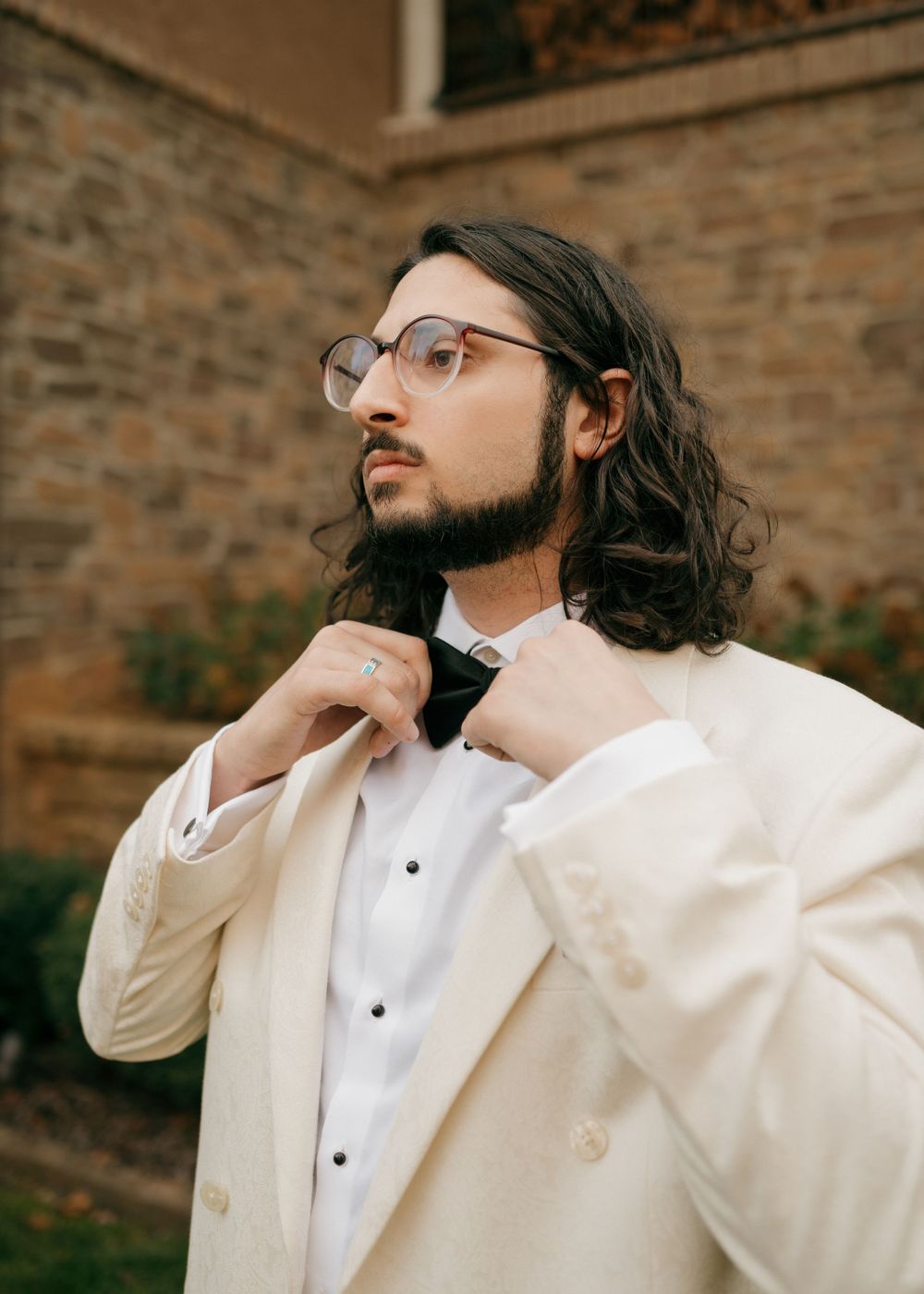Formal white double-breasted tuxedo jacket with bow tie and long dark hair at an outdoor wedding venue.