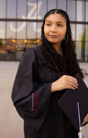 Graduate in a black academic gown poses outside a modern building with a diploma.
