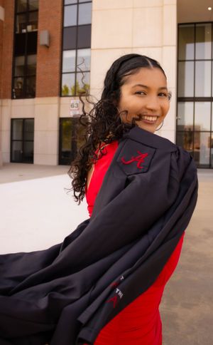 Graduate in red dress and black graduation gown posing outdoors with flowing curly hair.