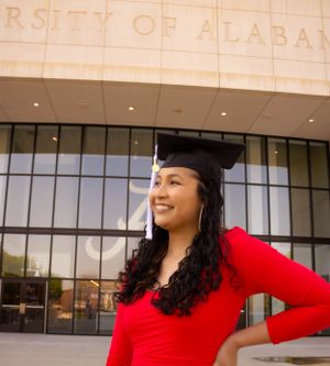 Graduate in red dress poses cheerfully outside the University of Alabama building.