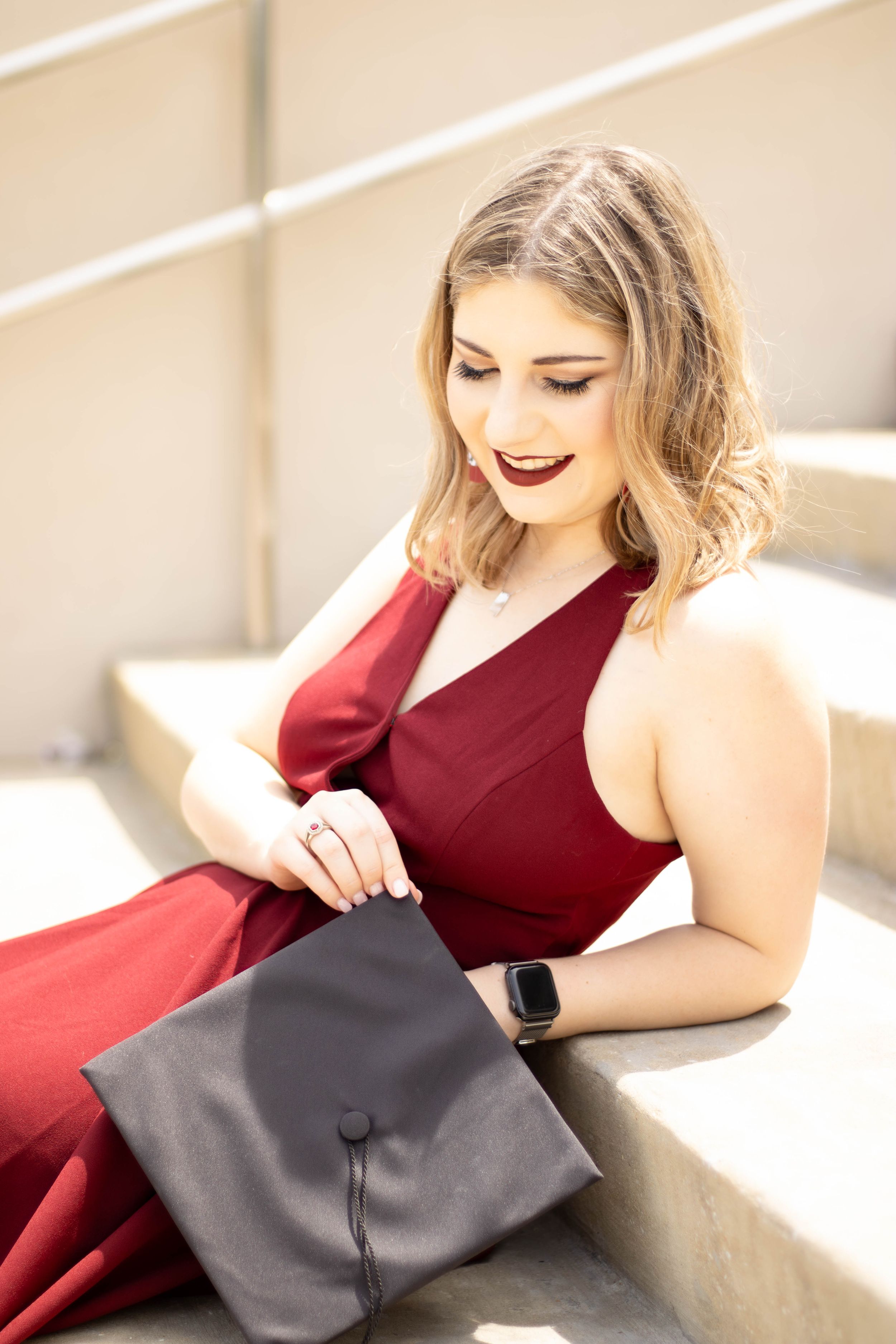 Graduate in red dress holding cap while sitting on concrete steps.