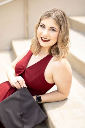 Portrait in burgundy dress holding graduation cap while reclining outdoors.