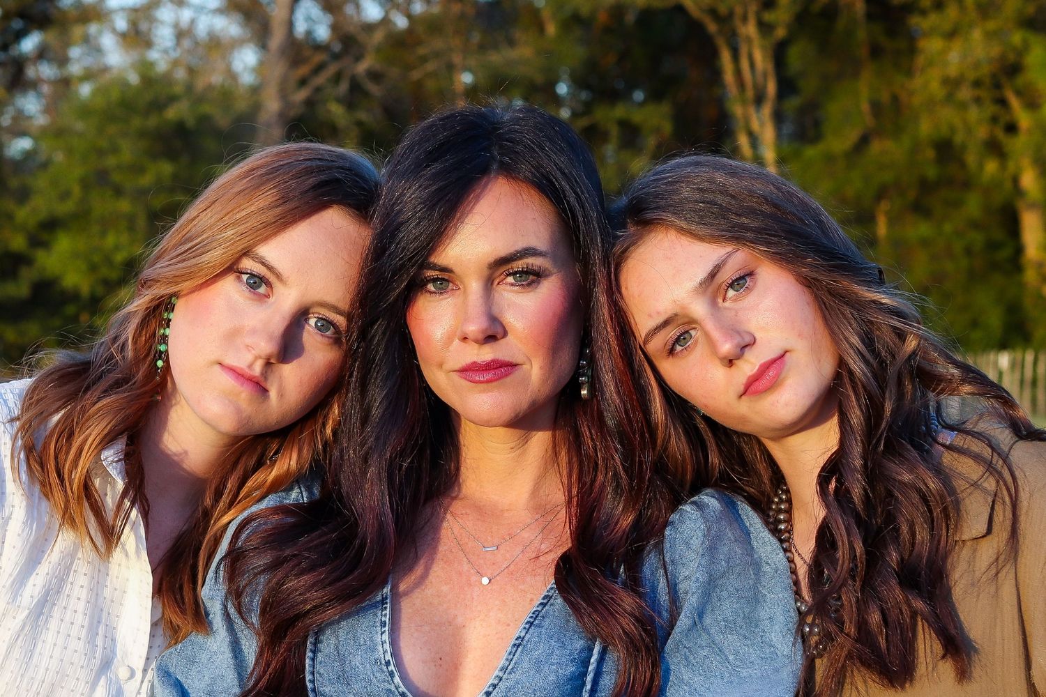 Three women with long brown hair posing closely together outdoors in warm golden light.