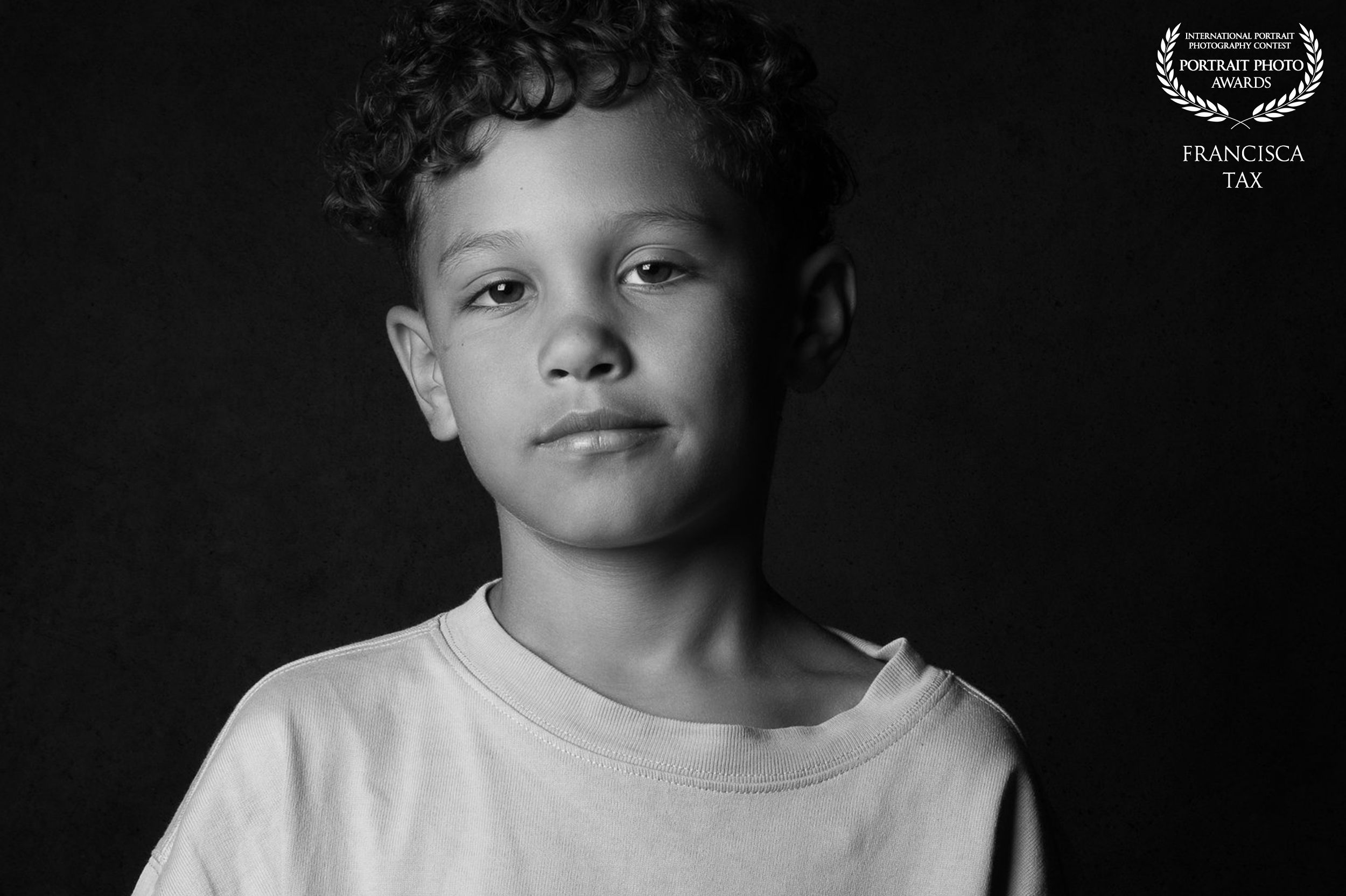 Black and white artistic portrait of a young child wearing a white t-shirt against a dark background.