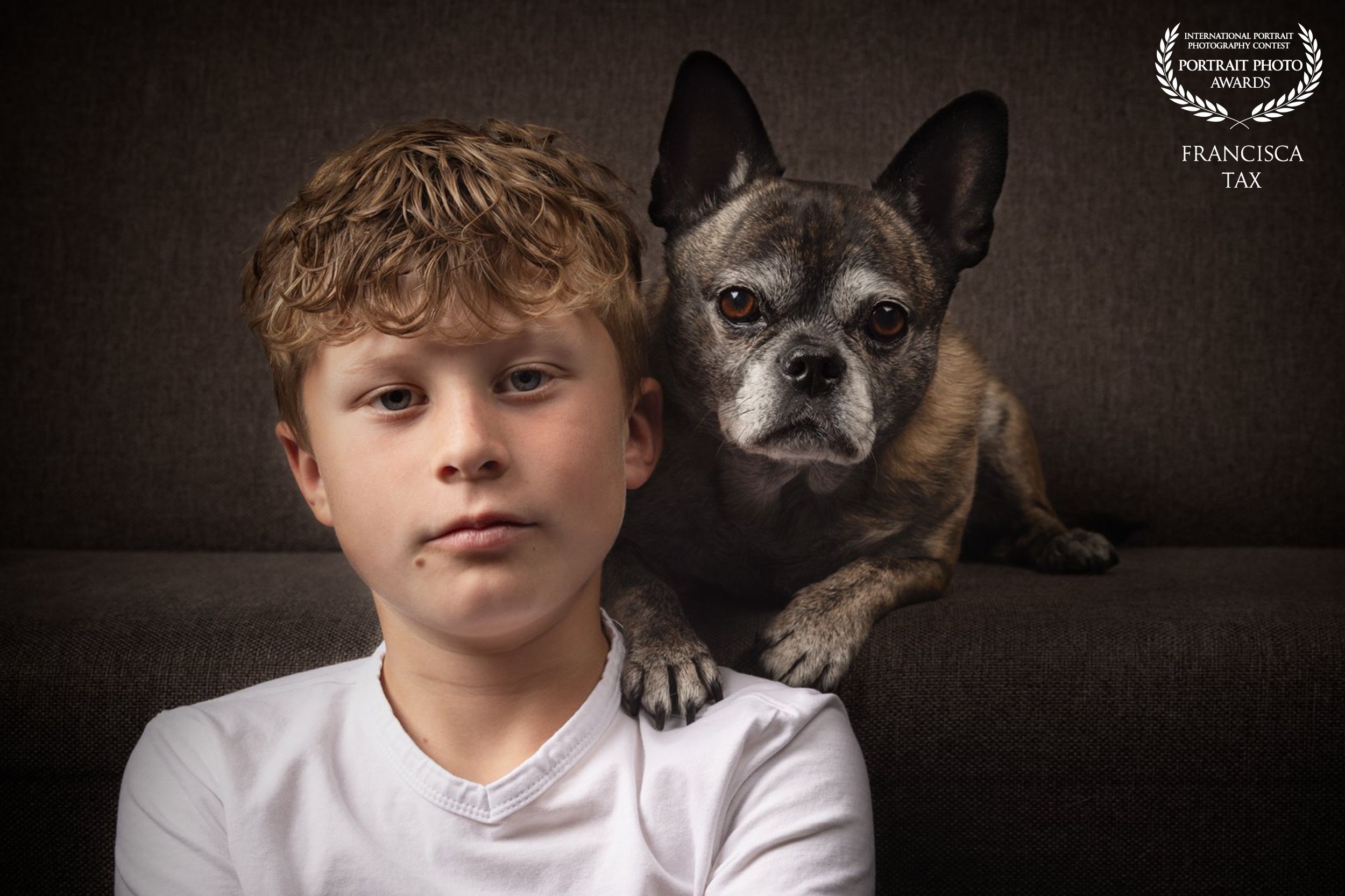 A child in a white shirt poses with a playful French Bulldog in a moody black background studio portrait.