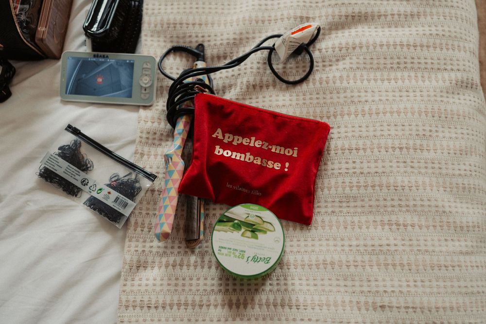 Wedding day essentials including jewelry and red pouch laid out on white textured bedding.
