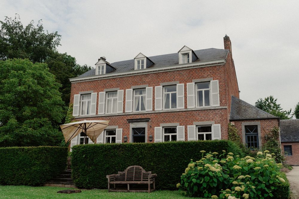 Elegant brick mansion with dormer windows and manicured hedges featuring a garden bench in the front yard.